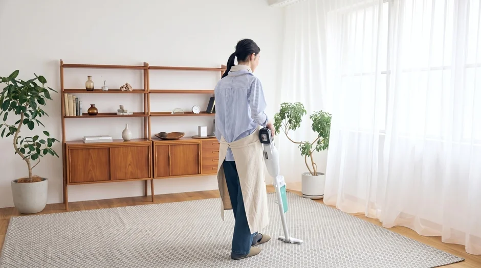 明るいリビングで、女性がコードレス掃除機を使ってカーペットを掃除しています。部屋には観葉植物や木製の棚があり、シンプルで清潔感のある空間が広がっています。
