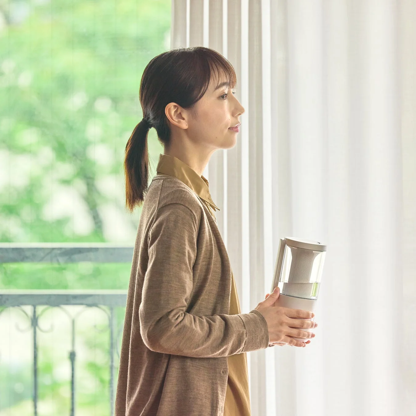 窓の外の緑を眺める女性が、白いポータブル家電のようなものを持っている様子